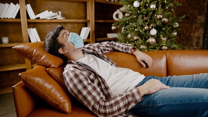 Young man in mask lies on sofa and stares at ceiling, suffering from loneliness and home isolation during Christmas holidays, coronavirus quarantine. Male suffering from boredom looking up covid 19
