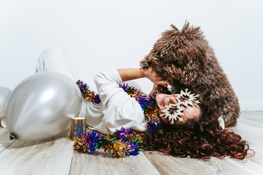 Brunette woman in pajamas lying down on the floor surrounded by balloons playing with her Spanish water dog at home on New Year's Eve. 2021 New Year's party at home concept