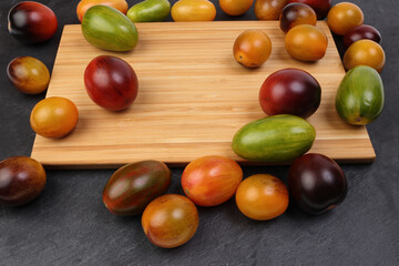 Red orange yellow green tomato mix variety on around wooden chopping board frame copy text space over black slate stone background