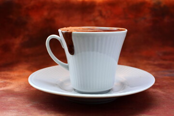Full to the brim white coffee cup on white porcelain saucer with strong aromatic black invigorating coffee, cocoa or hot chocolate flows out of seep in drop on the mug with foam on brown background