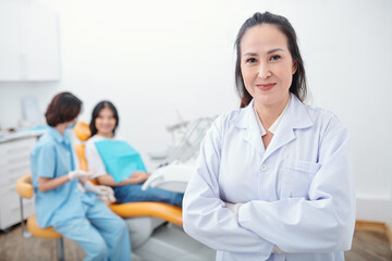 Portrait of confident smiling female Vietnamese dentist folding arms and looking at camera, her assistant talking to patient in background