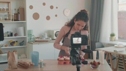 Tracking left medium of smiling Caucasian young woman standing at kitchen table filming for culinary vlog on semi-professional camera on tripod, talking, topping muffins with sprinkles