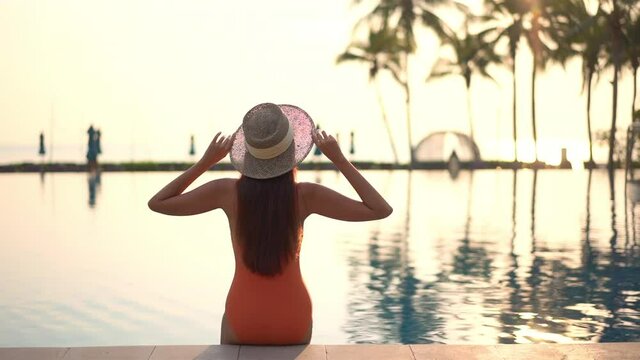 A woman with her back to the camera raises her hands into the air greeting the sun rising over a swimming pool and the ocean horizon beyond. 