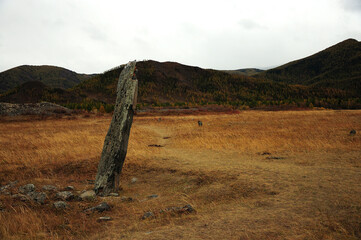 An ancient stone idol stands bent over in the autumn steppe and at the foot of high hills.