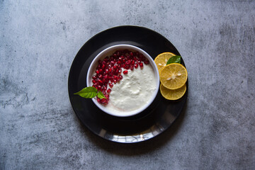 Pomegranate seeds on curd along with fresh fruit ingredients on a black plate.