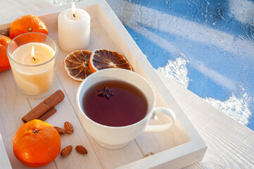 Winter tea at the frosty window. Tea with tangerines, cinnamon and nuts in a white cup on a white wooden tray.