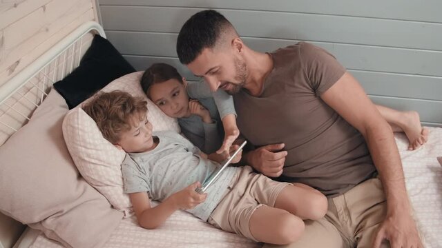 Medium shot of young father and cute little boy and girl relaxing on bed and using tablet