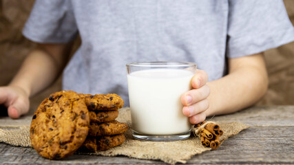 The child sits at a wooden table and holds a glass of milk, oatmeal cookies on the table.