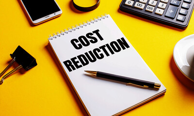 On a yellow background, a notebook with the text COST REDUCTION, coffee, calculator, telephone and a clip for papers. Business concept
