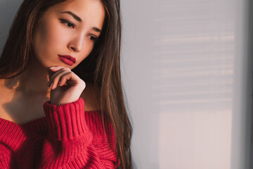 Sad young Asian woman sitting by the window, pensive, in the evening