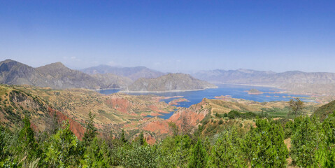 Beautiful panoramic view of Nurek dam lake on the Vakhsh river, second highest in world between Dushanbe and Khatlon regions in Tajikistan with trees foreground - obrazy, fototapety, plakaty