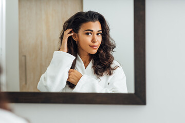 Beautiful female wearing bathrobe looking at a mirror and touching her hair