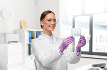 science, chemistry and people concept - happy smiling young female scientist in goggles with chemical sample or vaccine over laboratory background
