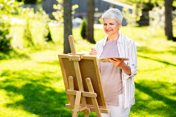 creativity, art and hobby concept - senior woman with easel and color palette painting outdoors