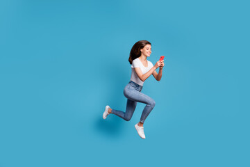 Obraz premium Full size profile side phot of brunette pretty girl run jump copyspace hold phone wear white t-shirt jeans isolated on blue color background