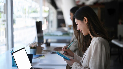 Side view of businesswoman working with computer laptop and taking note on notebook while sitting with her colleague at modern startup office.