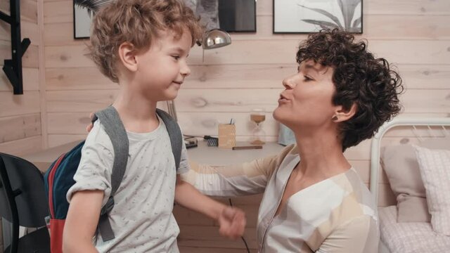 Medium shot of happy young mother with short hair putting schoolbag on cute little boy, then kissing him and waving goodbye as he is leaving for school
