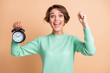 Photo of young beautiful girl excited happy smile fists hand win victory goal hold alarm clock isolated over beige color background