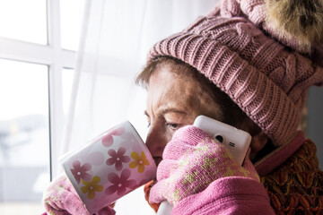 cold senior woman with cup looking out the window and talking on the mobile phone
