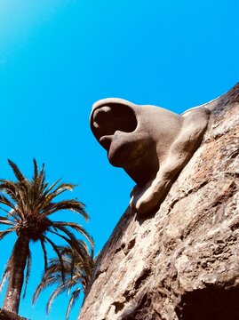 Low Angle View Of Sculpture Of An Animal Off The Wall Against Blue Sky With Palm Tree