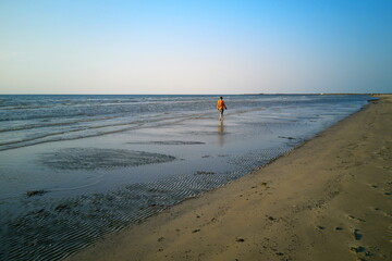 A lonely man in an orange jacket walks along the water along the shore.