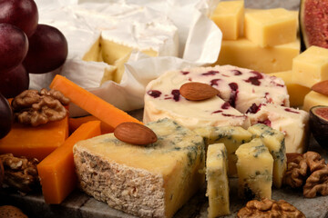 Cheese plate. Cheddar, Brie, Red Leicester, Wensleydale with sweetened dried cranberries, Blue Stilton, almond and walnut on a marble board with grape and figs on a black background.