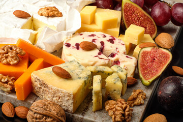 Cheese plate. Cheddar, Brie, Red Leicester, Wensleydale with sweetened dried cranberries, Blue Stilton, almond and walnut on a marble board with grape and figs on a black background.