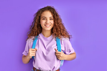 diligent shy caucasian school girl carrying school bag posing at camera, intelligent female teen isolated on purple background