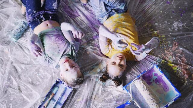 Children have fun in renovation room. Top view girl and boy smearing paints on skin and clothes fool around while lying near paint tool and tray on dirty, painted color floor covered protection film