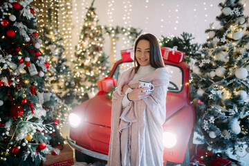 Young woman stands wrapped in plaid with cup of coffee in her hands near Christmas tree.