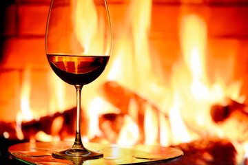 A glass with red wine against the background of a burning fireplace. Evening winter photo by the fireplace. The atmosphere of a cozy winter evening at home.