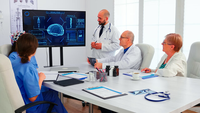 Doctor discussing about braing waves while is wearing headset with sensors. Monitor shows modern brain study while team of scientist adjusts the device working in hospital conference room - Powered by Adobe
