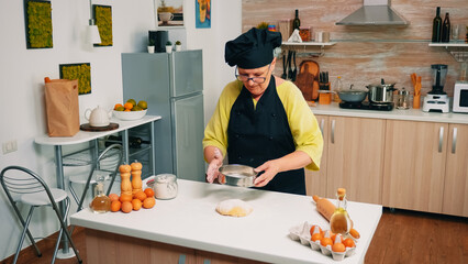 Woman baker sieving flour on bread dough with metallic sieve on table in kitchen. Happy elderly chef with uniform mixing sprinkling adding sifting spreading raw ingredients to baking traditional bread