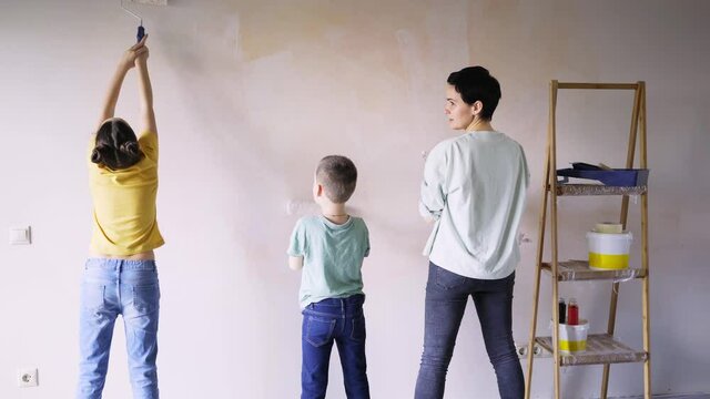 joyful mum with little son and daughter painting new apartment wall with rollers happiness standing near wooden ladder with tools and enjoying teamwork medium shot backside view