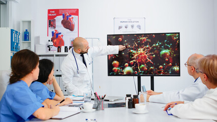 Specialist medic explaining virus development on digital screen in front of coworkers pointing on monitor in hospital office during brainstorming. Team of physicians analysing diagnosis of patients