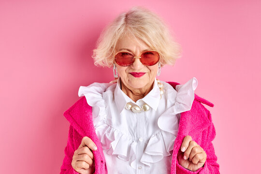 fancy aged lady in eyeglasses posing at camera wearing fashionable clothes, isolated over pink background