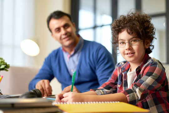 Portrait of teenaged latin boy in glasses smiling at camera, sitting at the desk together with his father and doing homework during learning at home