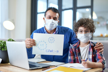 Safety. Middle aged hispanic father wearing mask sitting at the desk together with his son school boy and holding Covid 19 drawing during remote studying at home
