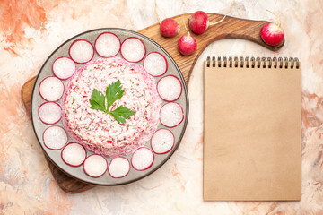 Delicious chicken salad with beet served with chopped red radishes on a gray plate on wooden cutting board and radishes next to notebook on mixed color background