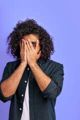 curly male is scared by something, close his face with hand, look at camera through one eye, isolated in studio