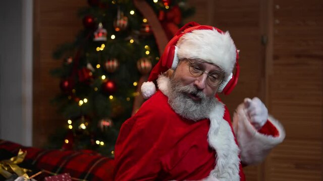 Portrait of Santa Claus dancing funny in big red headphones. Old man with beard in red suit and hat against the background of Christmas tree with toys and lights. Happy New Years evening. Slow motion.