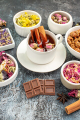 a cup of herbal tea with cinnamons and dry flowers and chocolate bars around it on grey background
