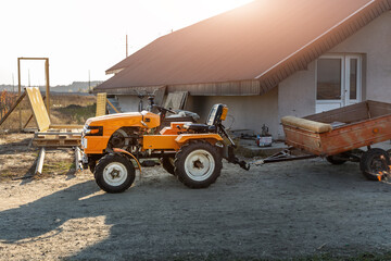 Small mini red modern orange new tractor with trailer standing near hangar building at farm countryside during sunset or sunrise. Small agricultural machinery. Rural country farmland scene background