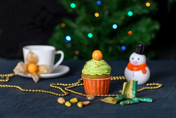 Pistachio cake with green cream and physalis on the table. In the background, a cup and saucer, a snowman, a Christmas tree with a garland and gold beads.