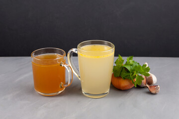 Two types of bone broth (yellow and brown) in transparent cups on a grey table. Dark background, selective focus, copy space. Fish and meat broth contain healthy collagen