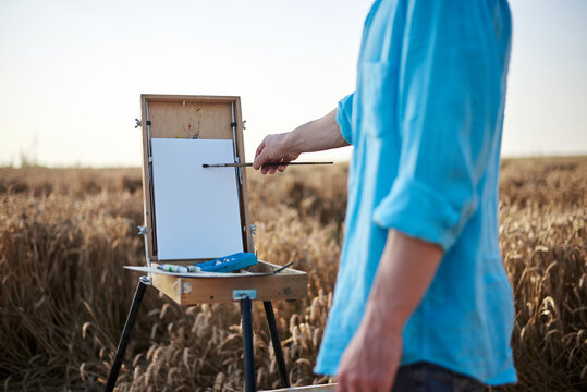 Close-up picture of hands of male artist, drawing on white canvas on wheat field in summer. Painting workshop in rural countryside. Artistic education concept. Outdoors leisure activities.