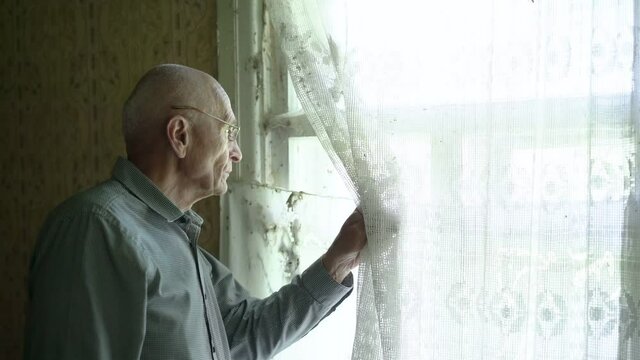 Aged senior man wears eyewear holds white curtain with hand and looks through window. Concept self isolation.