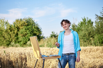 Young male artist wearing light blue shirt, drawing on canvas on sketchbook easel on wheat field. Painting workshop in rural countryside. Artistic education concept. Outdoors leisure activities.