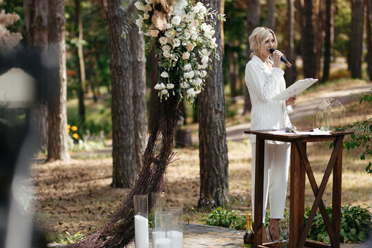 Master of wedding ceremony speeching on microphone on background wedding arch and trees.