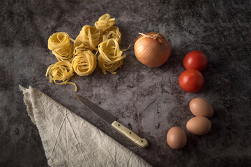 Fototapeta na wymiar Fresh onions, eggs, tomatoes and raw pasta with knife and table cloth on a stone table